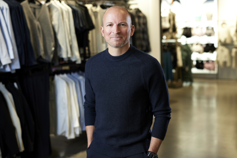 Smiling man in a navy sweater standing in a retail clothing store with shelves in the background
